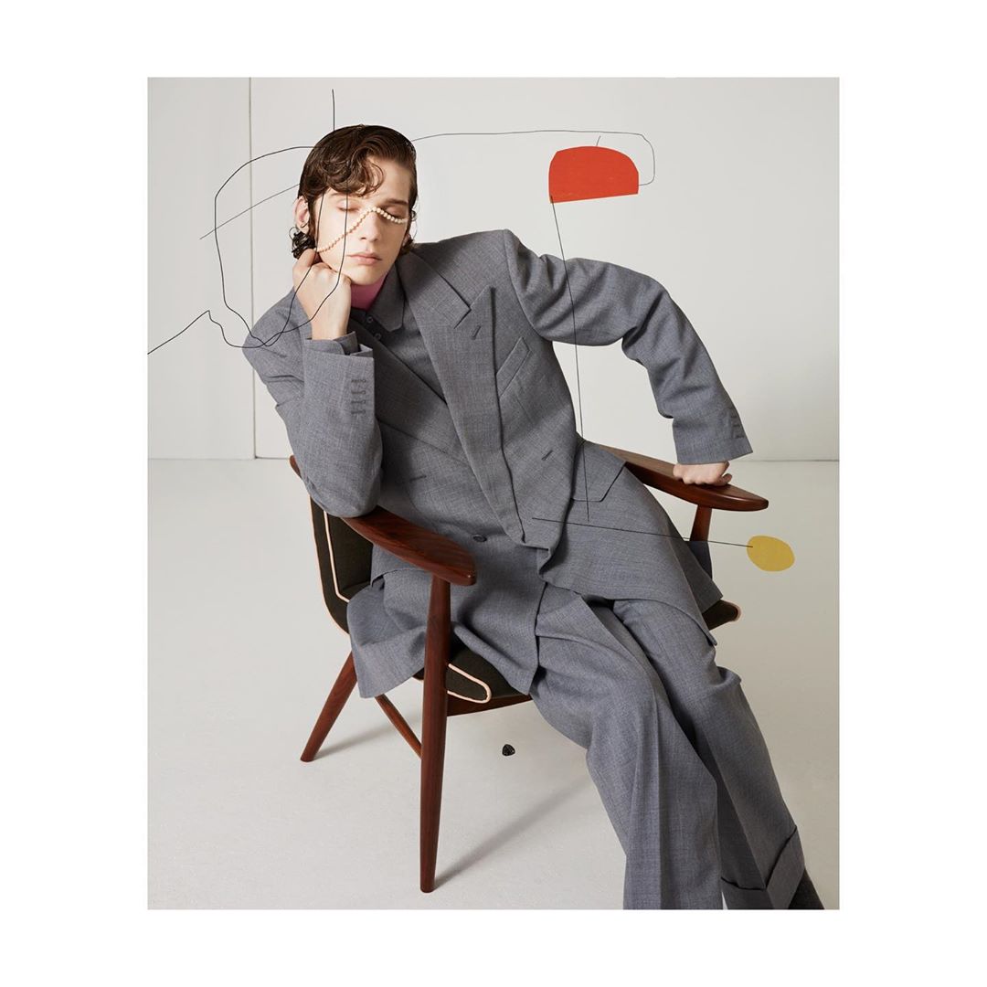 A young adult male model poses in a gray suit and pink shirt while seated in a wood-framed chair with an abstract painting behind him.