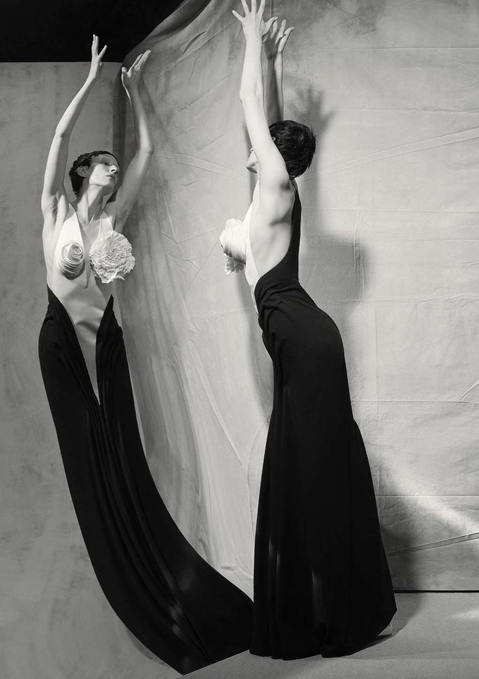 Two women in elegant black gowns strike dramatic poses against a textured backdrop, captured in a black and white studio fashion shot.