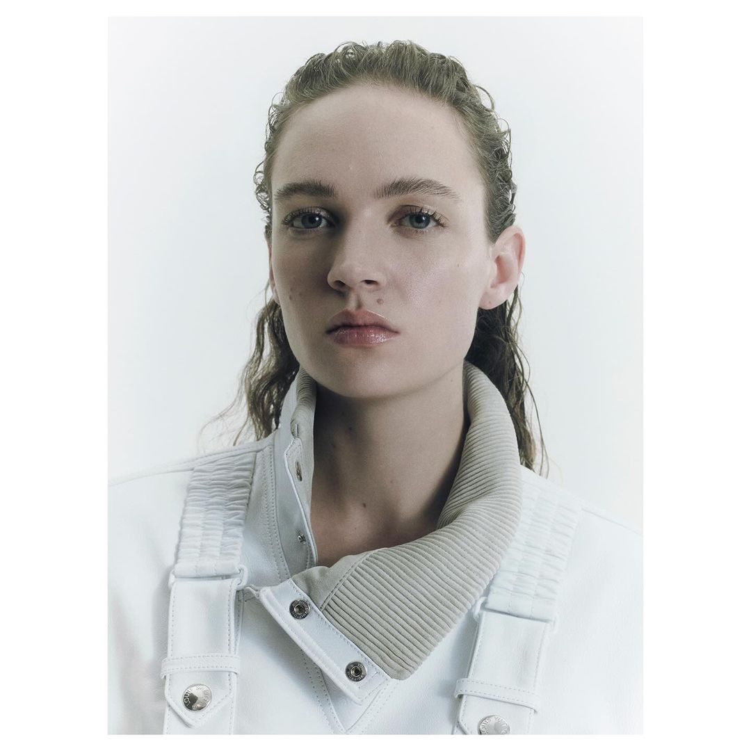 Close-up portrait of a young woman with damp hair wearing a white jacket with a light tan ribbed collar against a bright white backdrop.