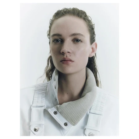 Close-up portrait of a young woman with damp hair wearing a white jacket with a light tan ribbed collar against a bright white backdrop.