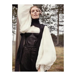 Model in voluminous white bishop-sleeve blouse and black structured top standing outdoors among pine trees.