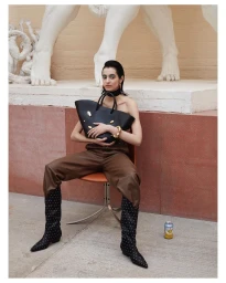 Model with slicked-back dark hair holding a large black leather handbag while seated on an orange chair outdoors