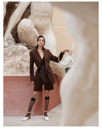 Model in a brown blazer holding a white handbag standing before a large classical stone sculpture