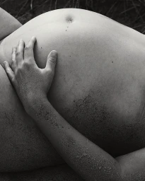 Monochrome close-up of a pregnant belly with a hand resting gently on it, dusted with sand.