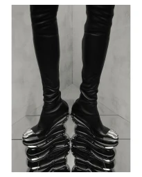 Close-up of black leather thigh-high boots with metallic sculptural heels against a reflective surface and textured concrete corner wall.