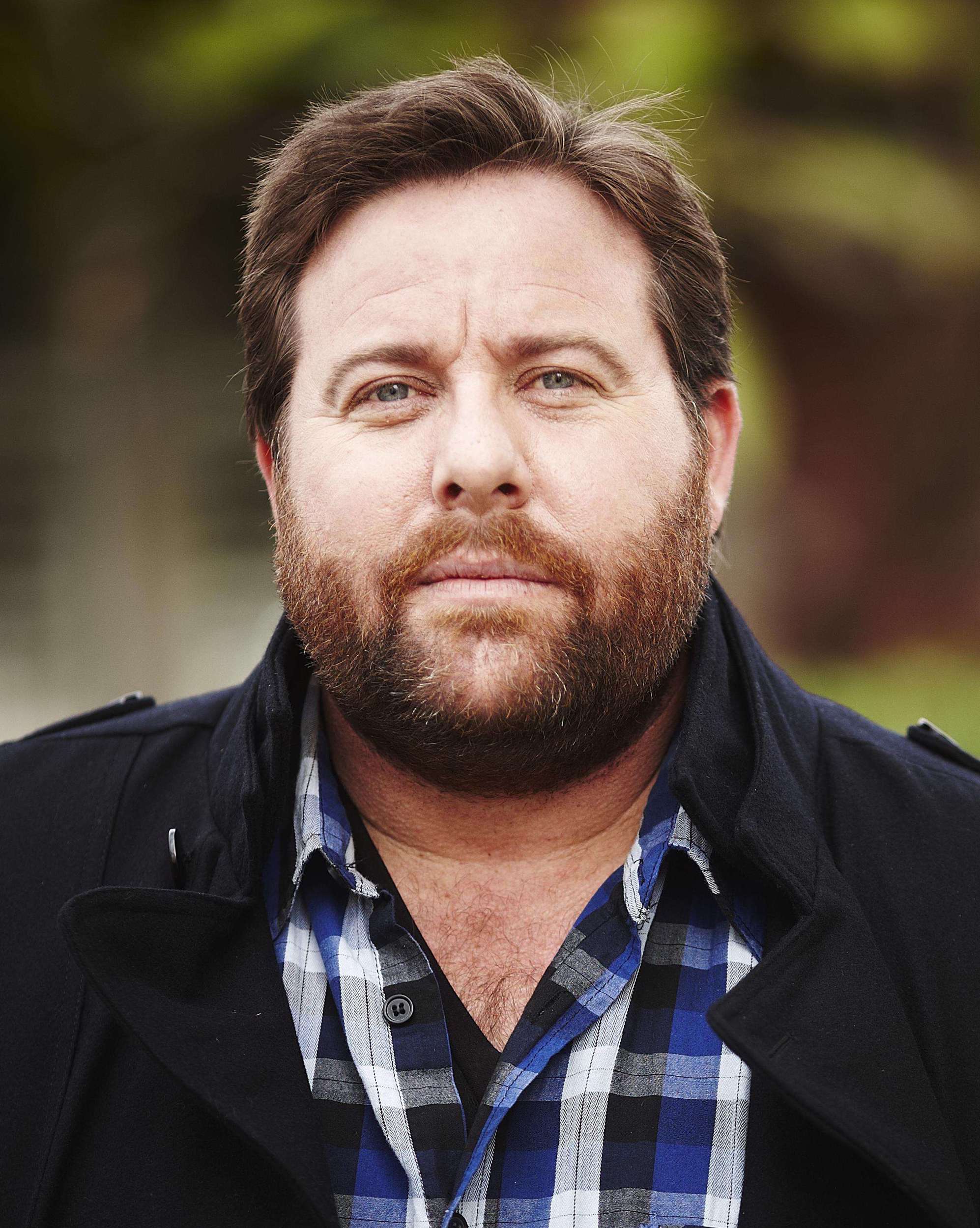 Portrait of a middle-aged man with a beard, wearing a blue plaid shirt and a dark coat against a blurred green background outdoors.