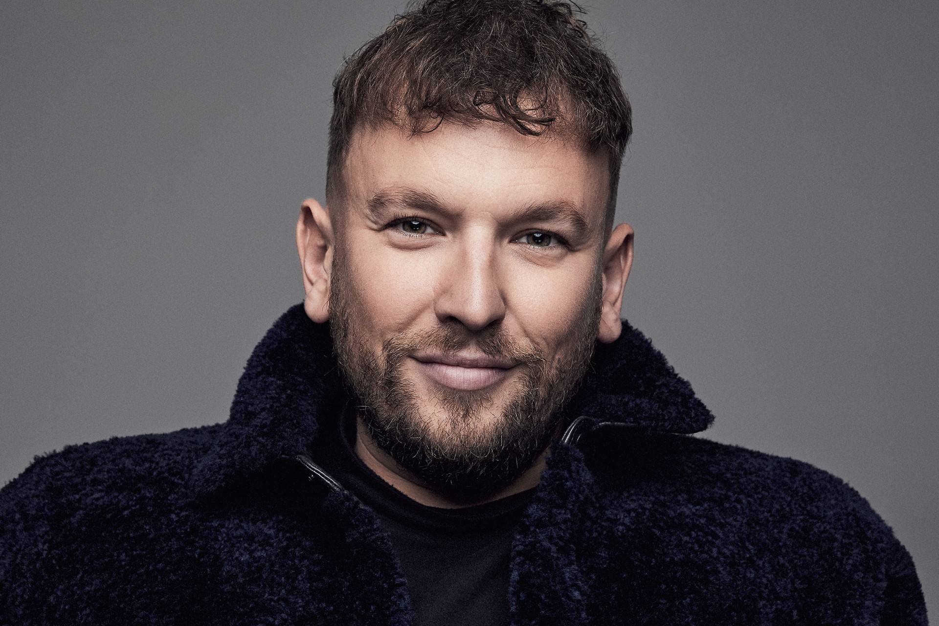 Portrait of an adult male with a beard, wearing a dark fuzzy jacket. The subject has a friendly smile and is set against a grey backdrop.