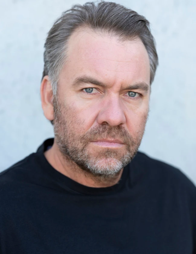 Portrait of a middle-aged man with short grey hair and a stubble beard wearing a black t-shirt against a plain light wall.