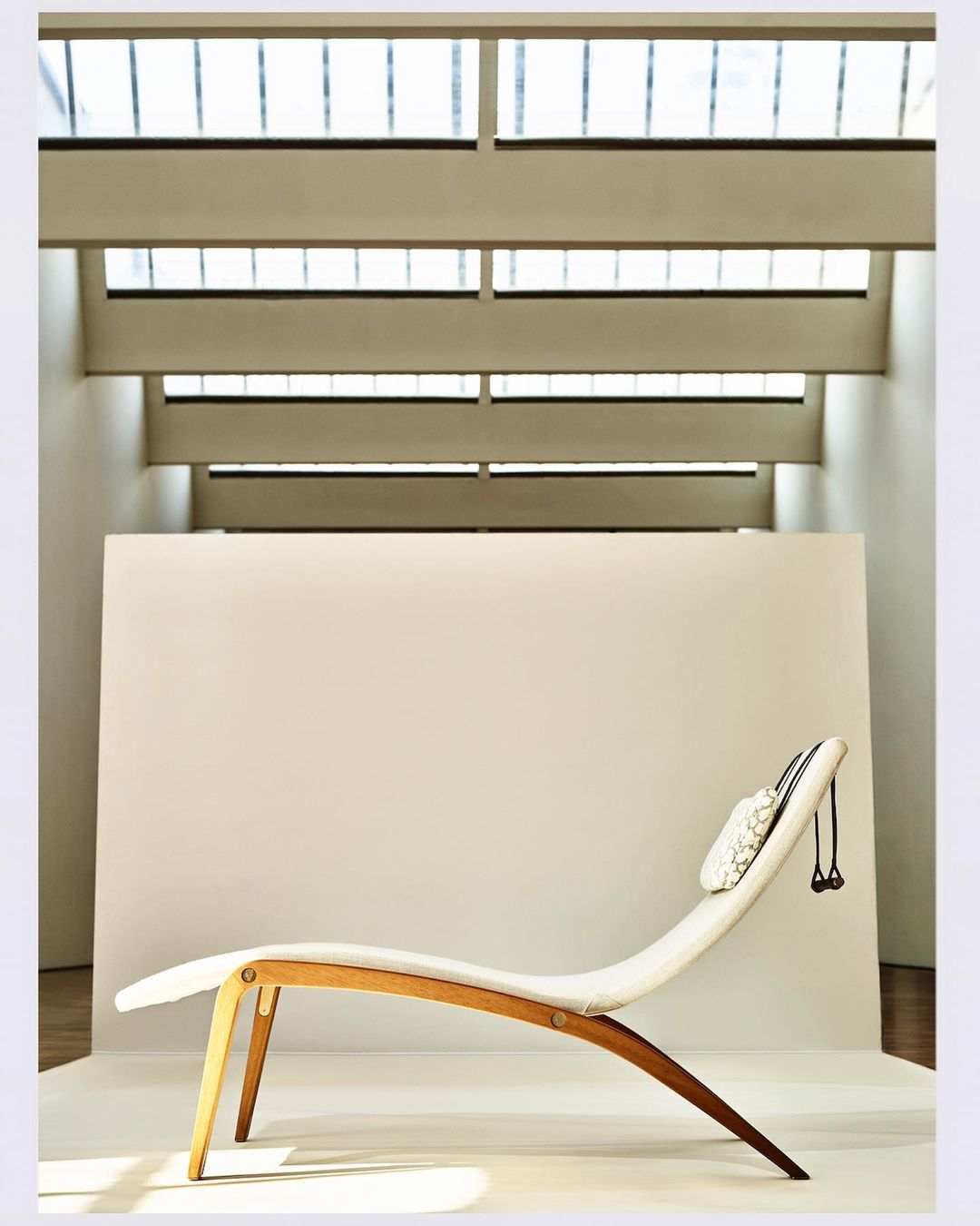 Minimalist interior shot of a wooden and white fabric chaise lounge with a neck pillow, set against a plain white backdrop with window light.