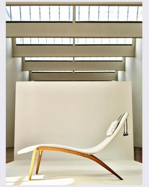 Minimalist interior shot of a wooden and white fabric chaise lounge with a neck pillow, set against a plain white backdrop with window light.