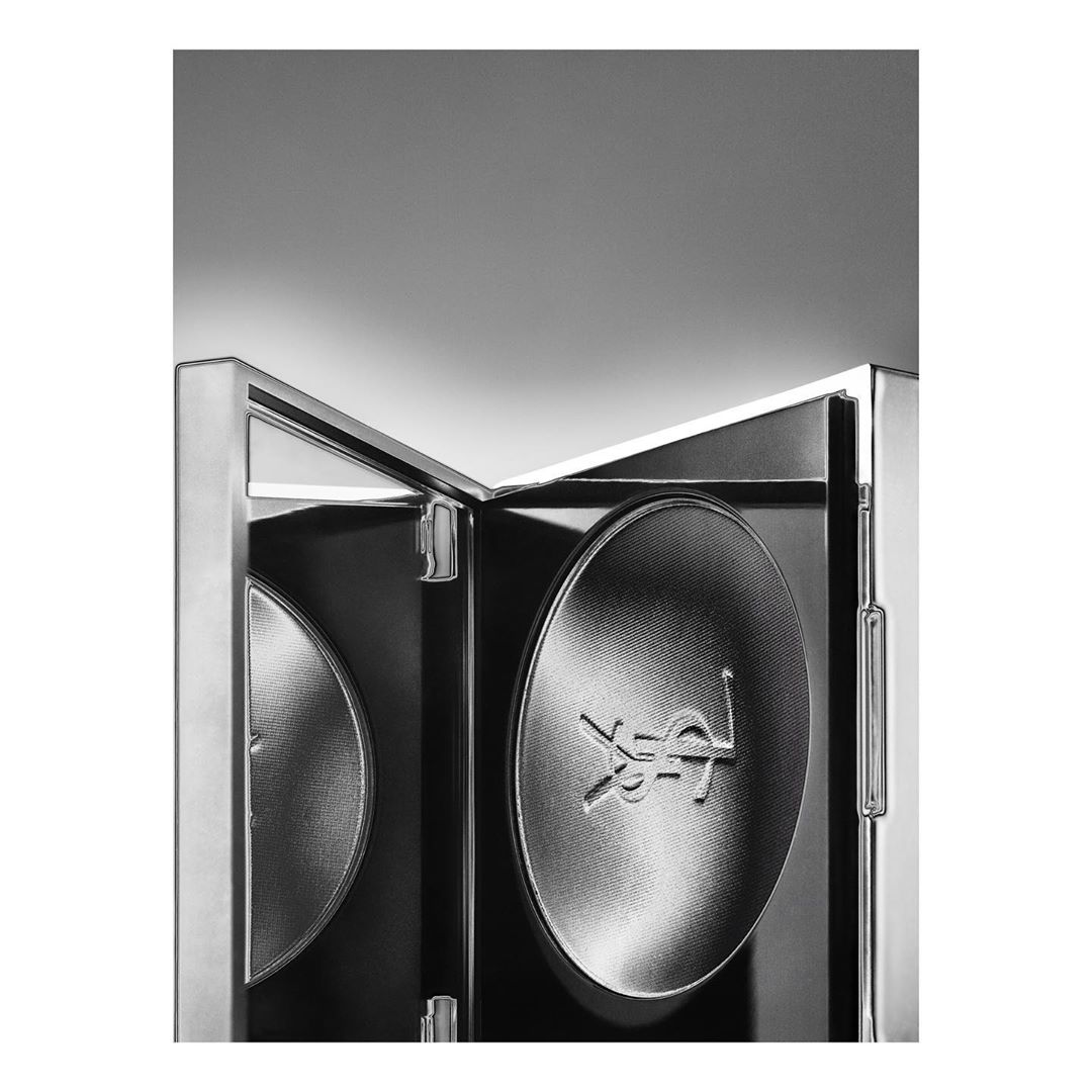 Monochrome beauty shot of an open silver compact case with two round powder pans, one embossed with the YSL logo, and a gray backdrop.