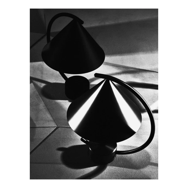 Monochrome photograph of two lamps with cone-shaped shades, casting geometric shadows on a textured surface, creating stark contrast.