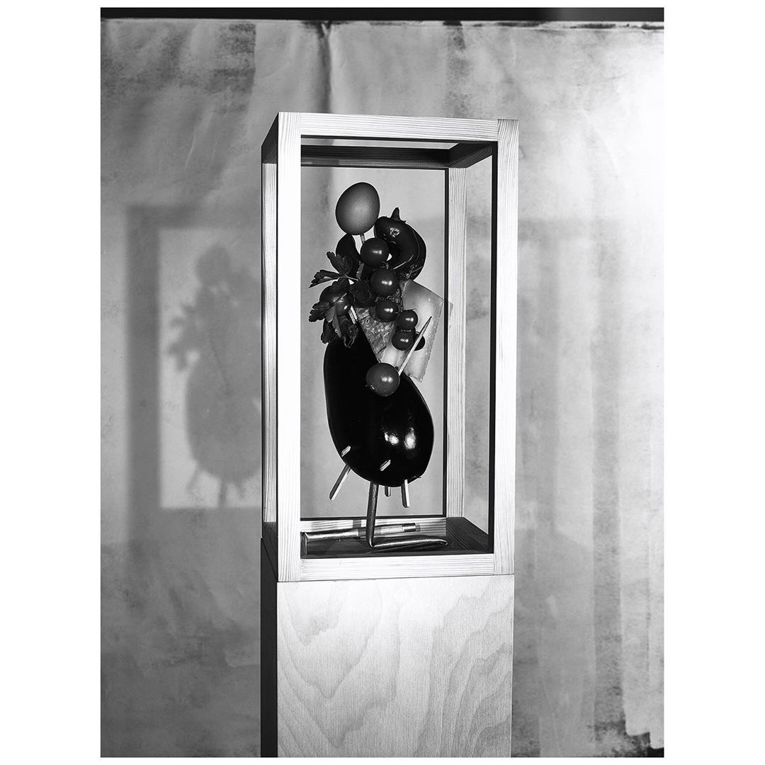 Still life sculpture in a wooden box on a stand featuring eggplant, grapes, a peach, and leaves against a blurred background.