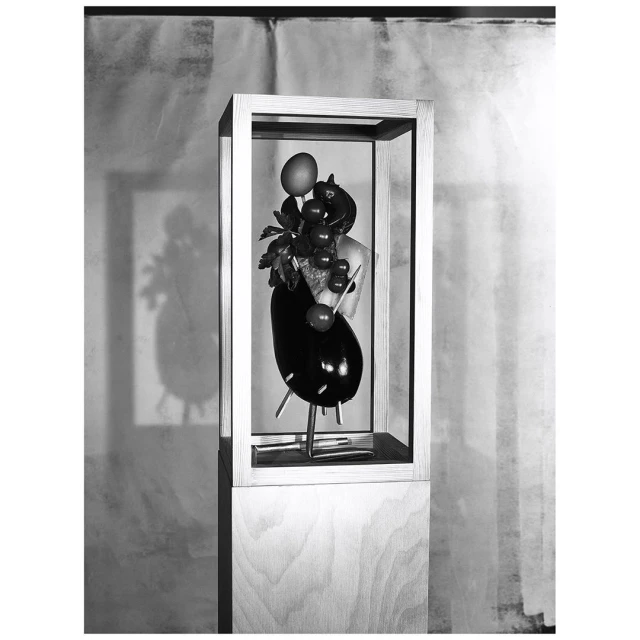 Still life sculpture in a wooden box on a stand featuring eggplant, grapes, a peach, and leaves against a blurred background.