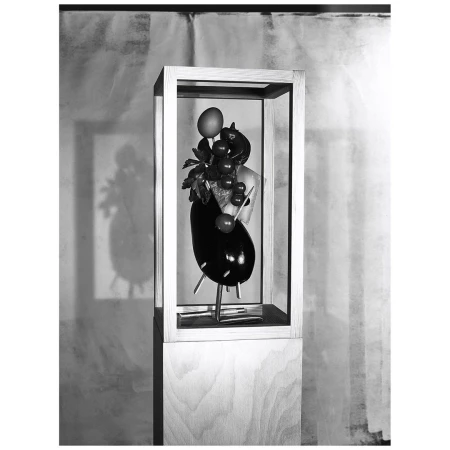 Still life sculpture in a wooden box on a stand featuring eggplant, grapes, a peach, and leaves against a blurred background.