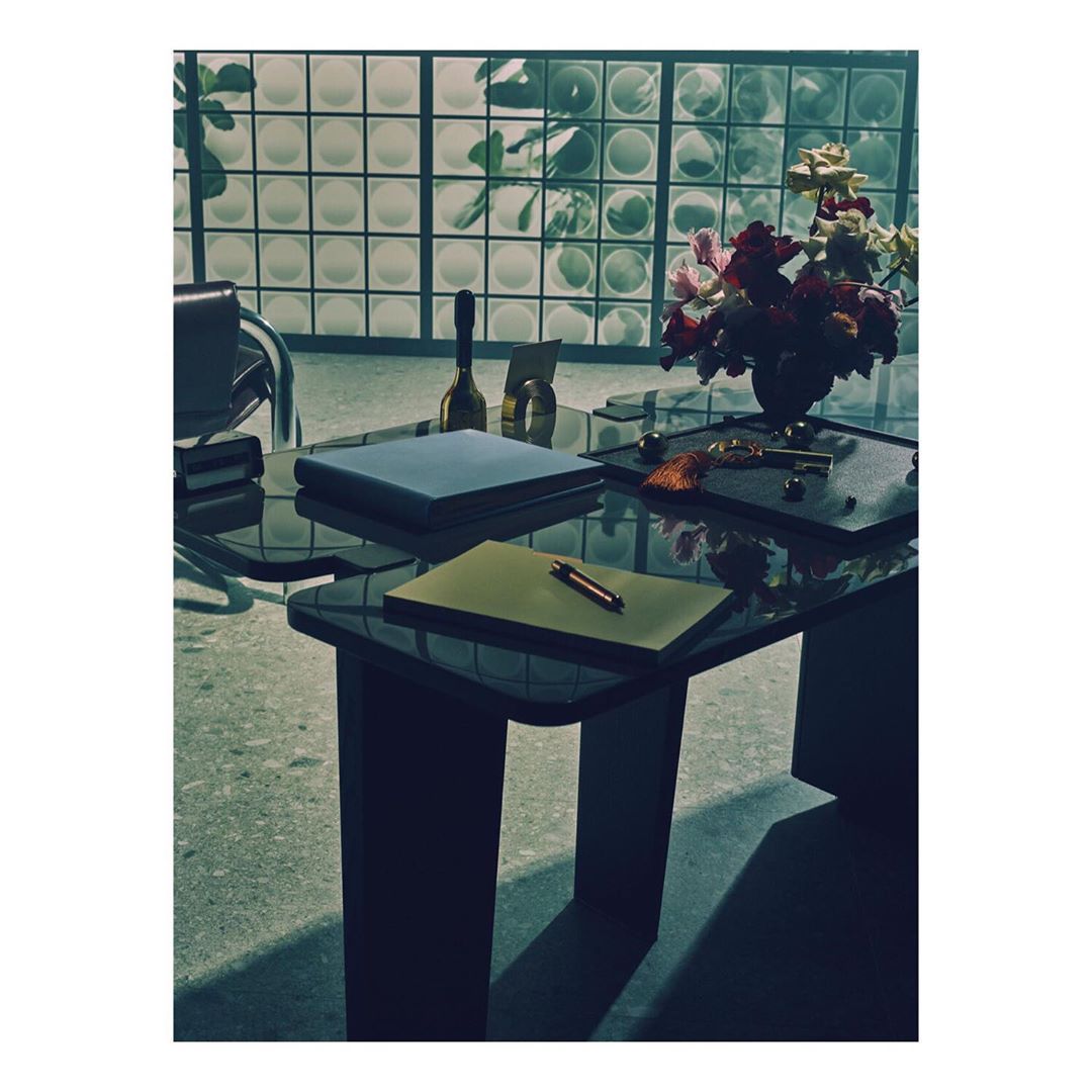 Interior shot of a dark glass-topped desk with office objects and a flower arrangement, set against a bubbly glass wall and flooring.