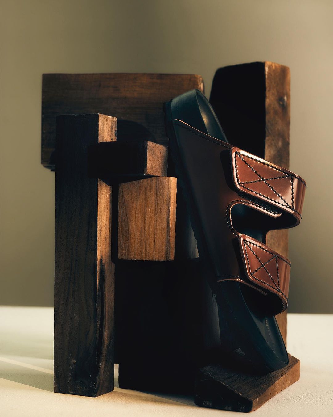 Studio shot of a brown leather sandal displayed with geometric dark brown wooden blocks on a beige surface with a plain beige background.