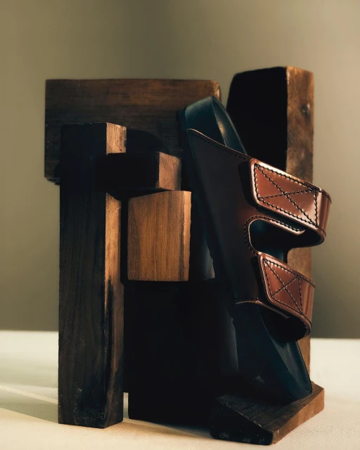 Studio shot of a brown leather sandal displayed with geometric dark brown wooden blocks on a beige surface with a plain beige background.