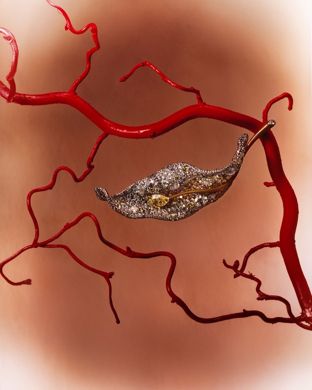 Leaf-shaped jewelry brooch encrusted with diamonds and yellow gems, attached to a bright red branch on a peach-colored gradient background.