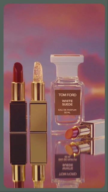 Cosmetic still life: Tom Ford lipsticks and White Suede eau de parfum on a reflective surface, with a pink and orange clouded background.