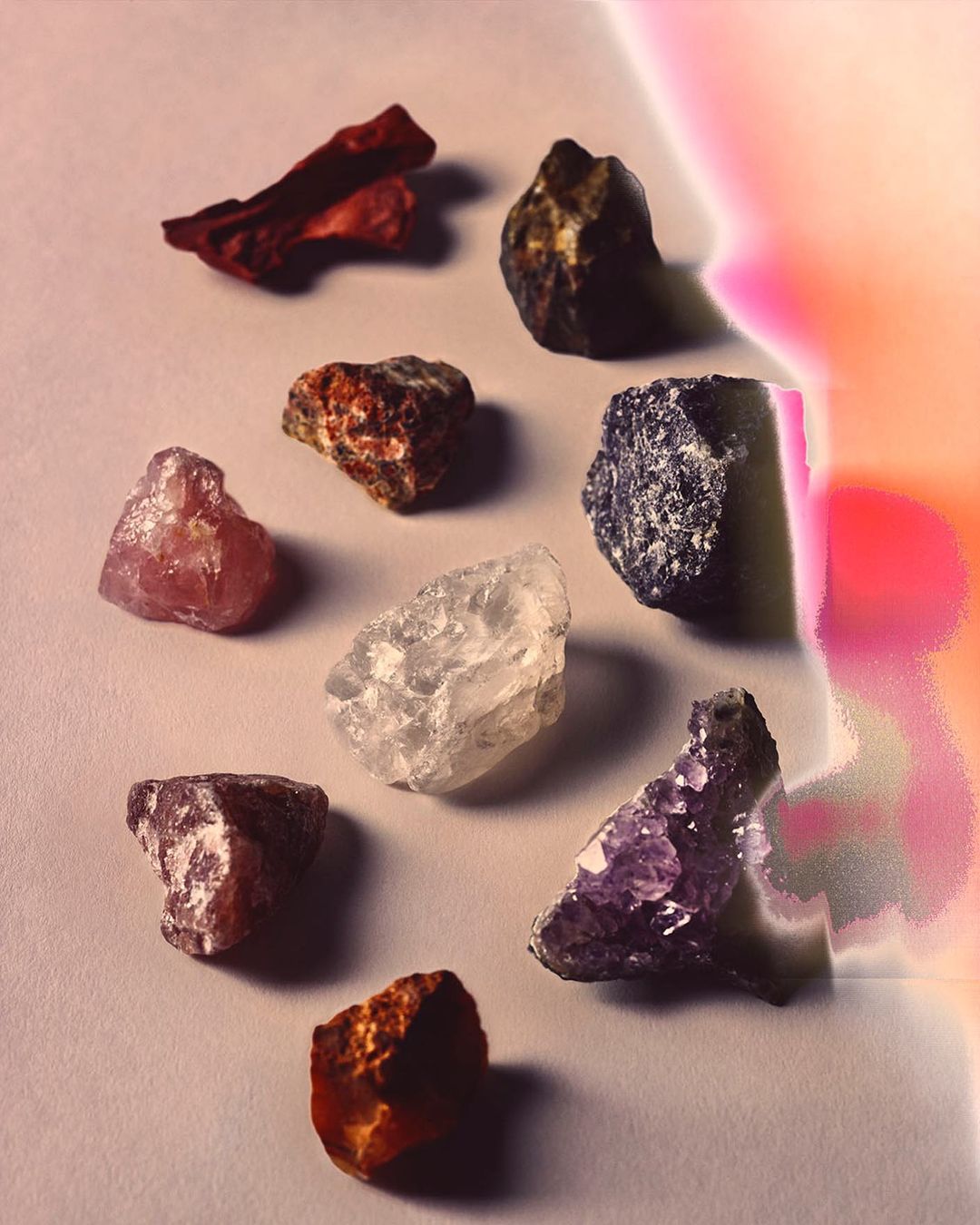 Macro image showcasing a collection of raw mineral specimens with varying colors and textures arranged on a white surface with soft pink light.