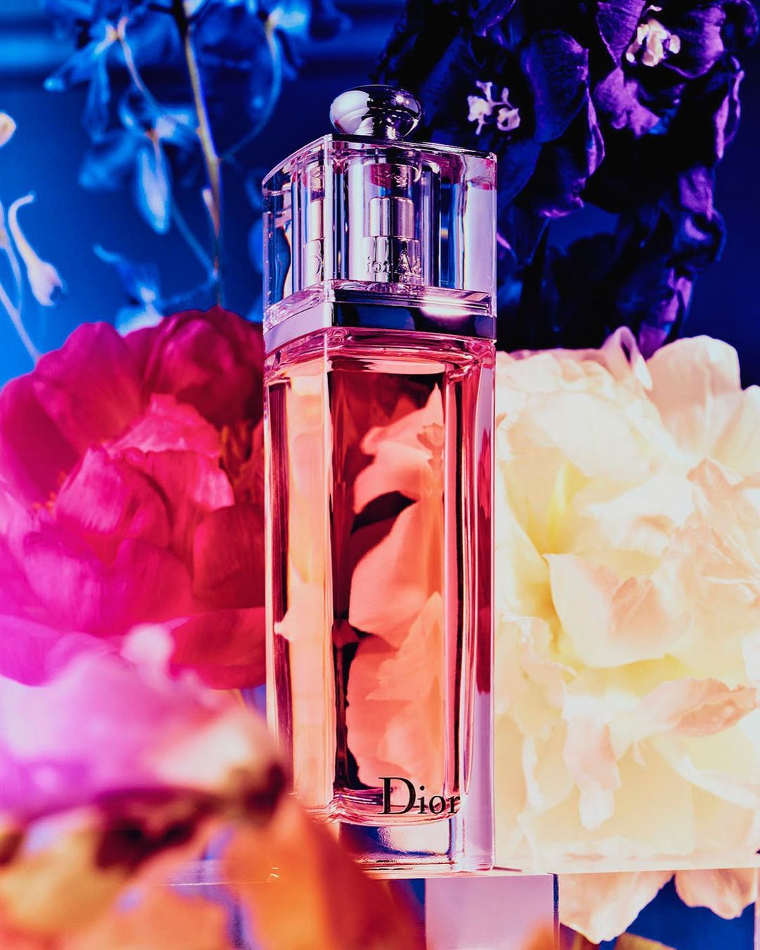Close-up product shot of Dior perfume bottle with floral background in pink, blue, and white. Glass and metal details visible.