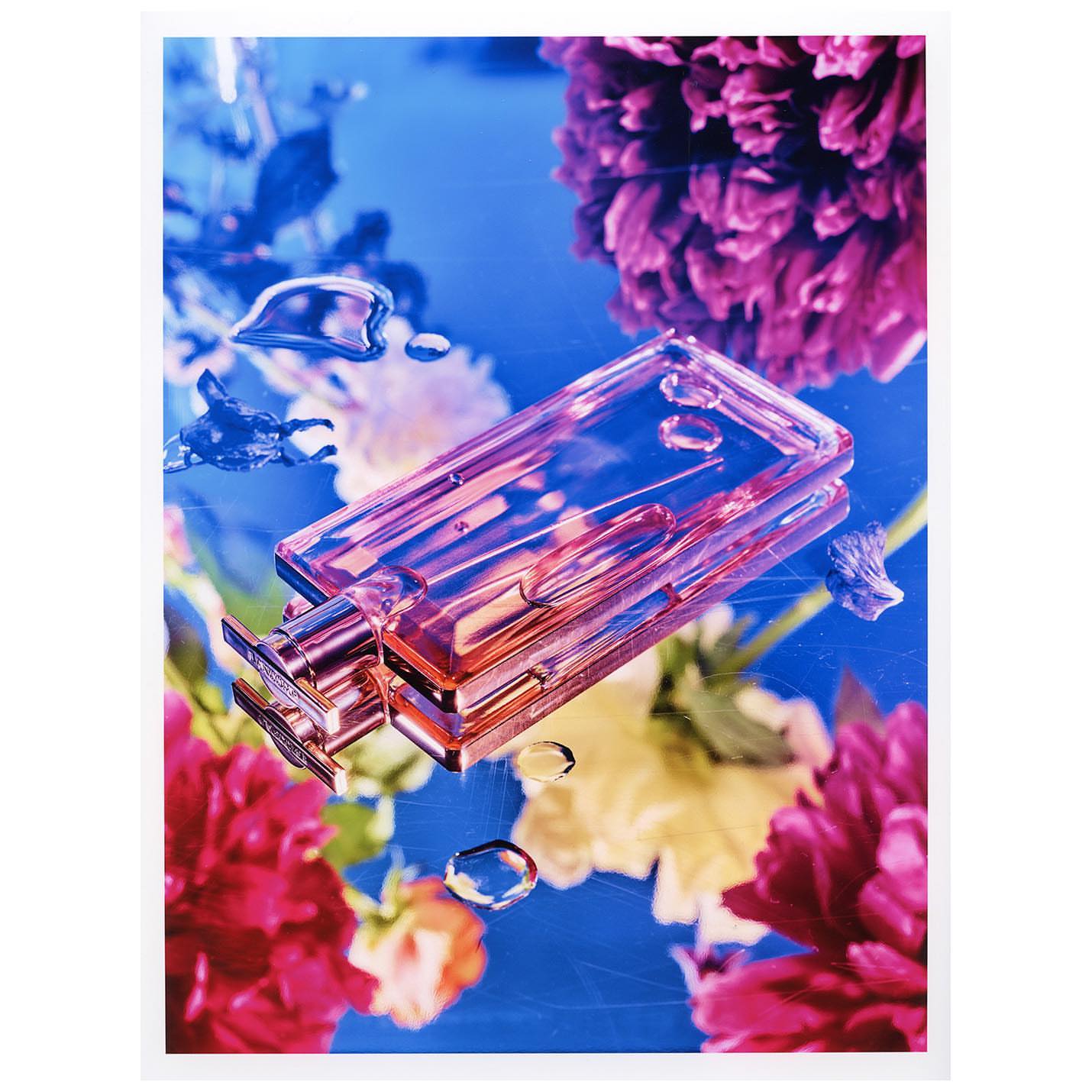 Rectangular glass perfume bottle resting on a reflective blue surface surrounded by vibrant pink floral blossoms and water droplets.
