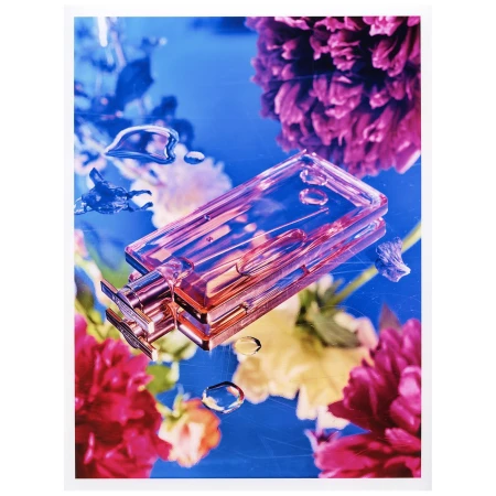 Rectangular glass perfume bottle resting on a reflective blue surface surrounded by vibrant pink floral blossoms and water droplets.