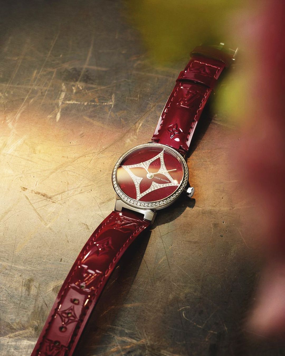 Close-up of a Louis Vuitton Tambour Moon Divine watch on a textured metallic surface showcasing a red leather strap and diamond details.