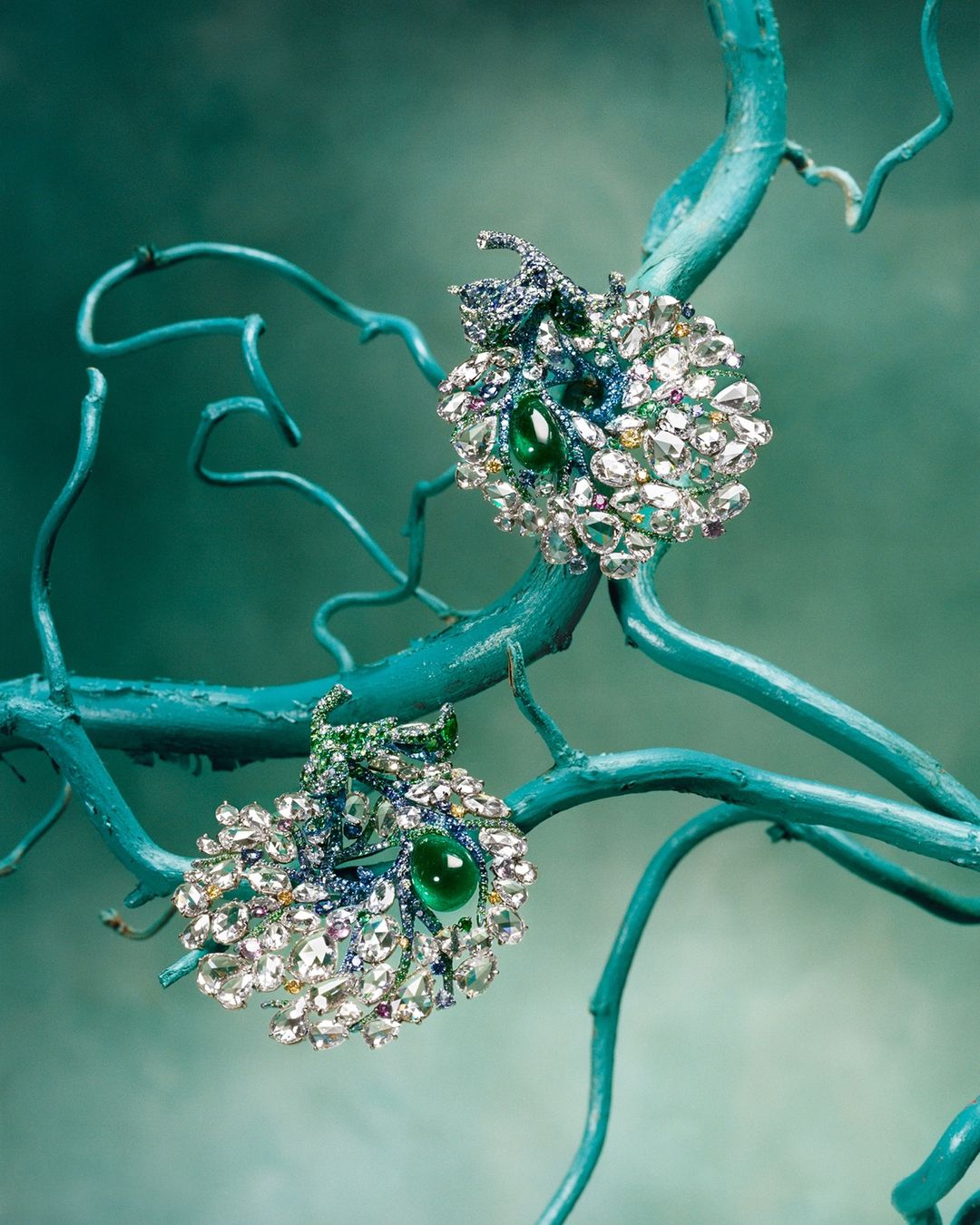 Elegant pair of decorative earrings featuring emeralds and sparkling crystals, set against a turquoise painted branch and background.