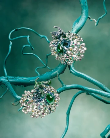 Elegant pair of decorative earrings featuring emeralds and sparkling crystals, set against a turquoise painted branch and background.