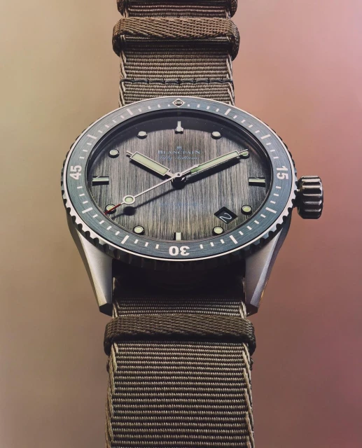 Close-up of a Blancpain watch with a textured gray dial, silver case, and an olive green nylon strap displayed on a neutral backdrop.