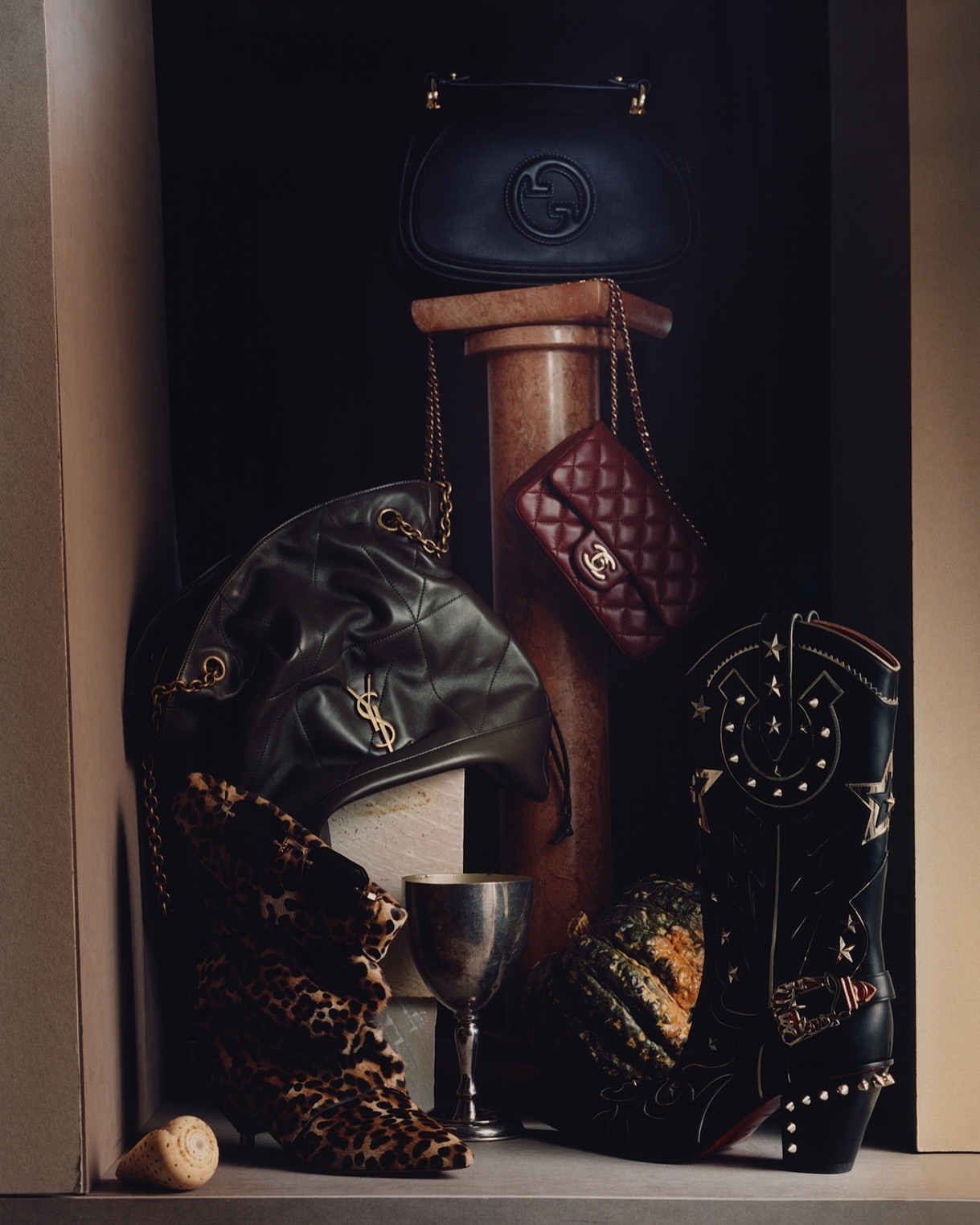 Still life with designer handbags from Gucci, Chanel, and Yves Saint Laurent, leopard print booties, and a cowboy boot on a dark backdrop.