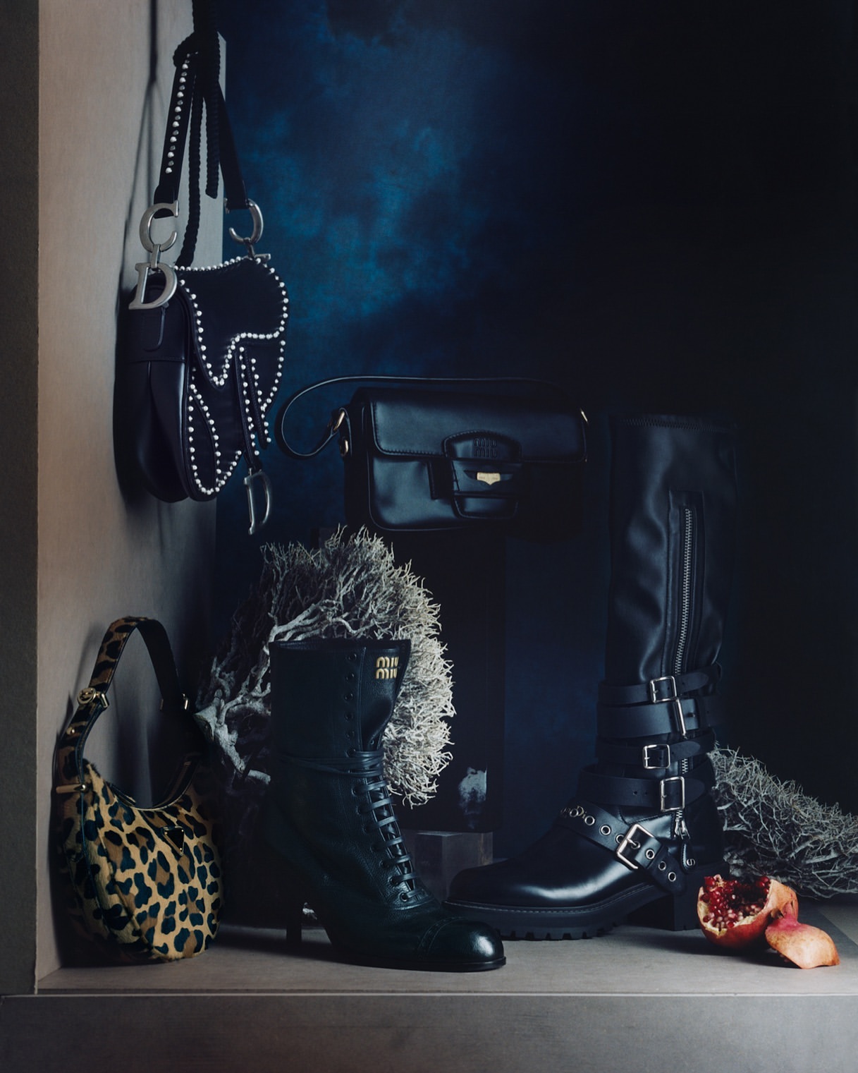Fashion still life featuring designer handbags and boots in a dark, moody setting with dramatic lighting and luxe textures, and a sliced pomegranate.