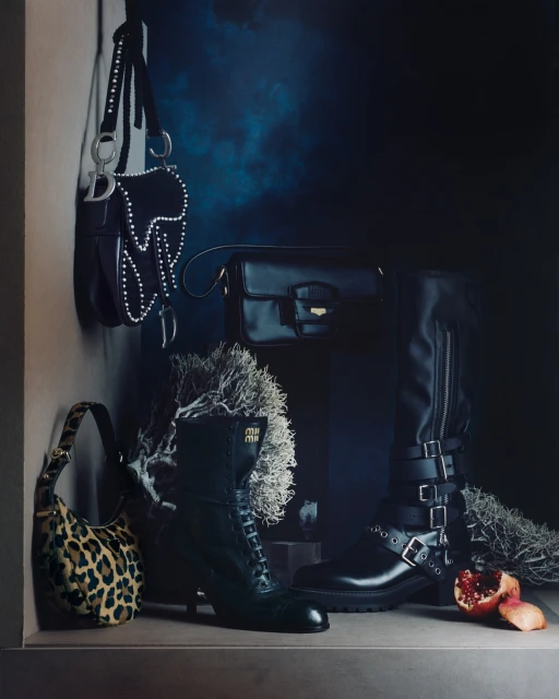 Fashion still life featuring designer handbags and boots in a dark, moody setting with dramatic lighting and luxe textures, and a sliced pomegranate.