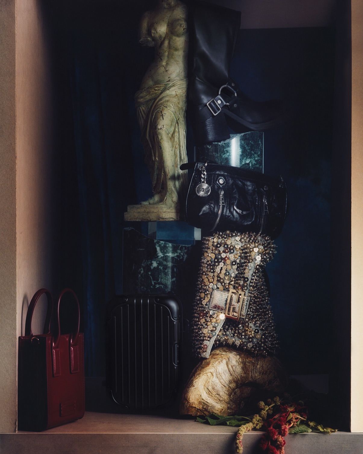 Still life photograph of stacked bags, a statue, suitcase, and floral elements in subdued lighting, conveying an artistic, curated composition.