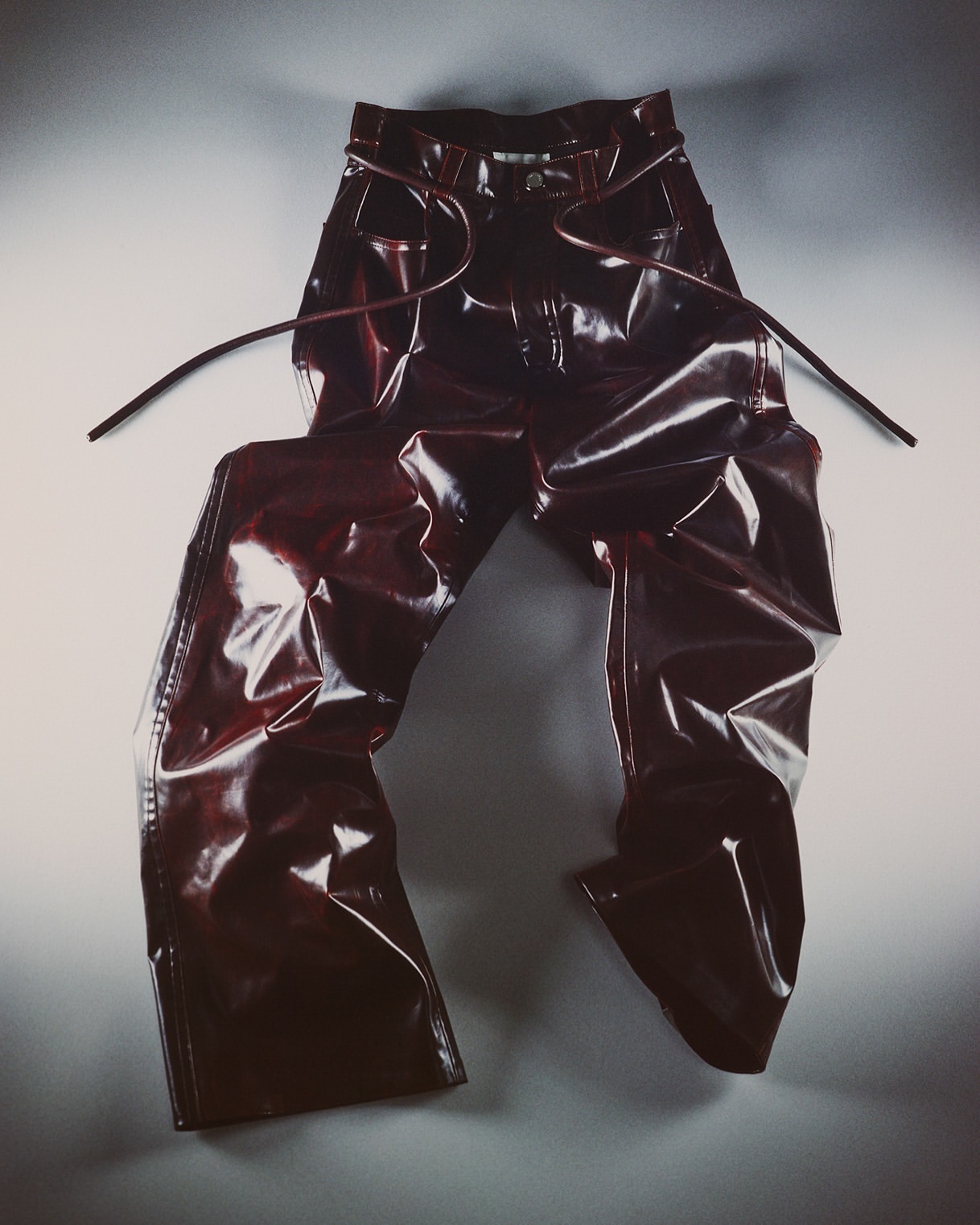 Shiny red vinyl pants with tied openings are displayed on a light gray surface in a product shot against a light gray backdrop. 