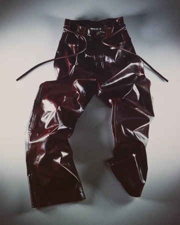 Shiny red vinyl pants with tied openings are displayed on a light gray surface in a product shot against a light gray backdrop. 