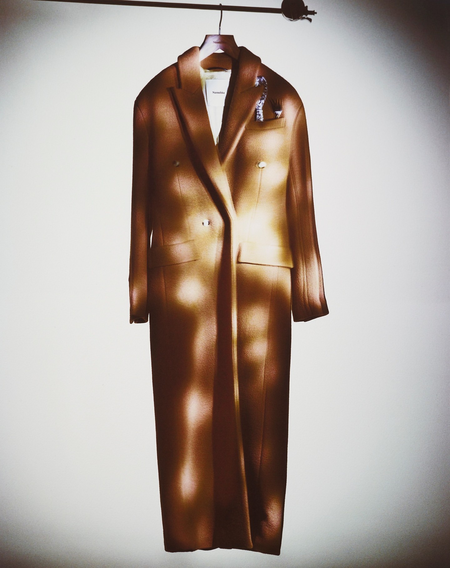 Studio shot of an elegant tan overcoat on a hanger against a white wall with dappled lighting for a fashion or retail display.