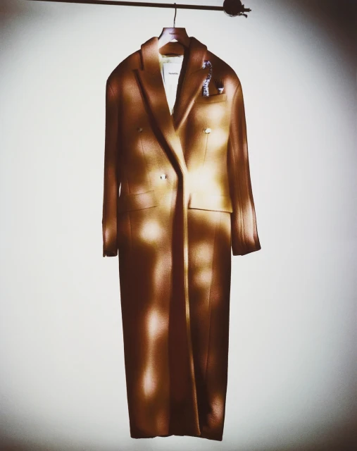 Studio shot of an elegant tan overcoat on a hanger against a white wall with dappled lighting for a fashion or retail display.