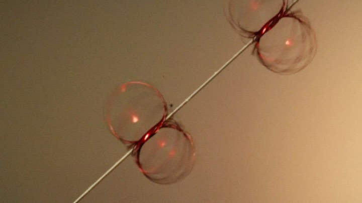Abstract close-up of bubbles connected to a thin wire, suspended in the air with a neutral backdrop. Studio lighting illuminates the scene.