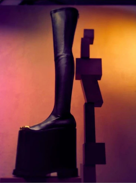 Black platform thigh-high boot stands next to a geometric sculpture against a warm orange and pink backdrop in a studio setting.