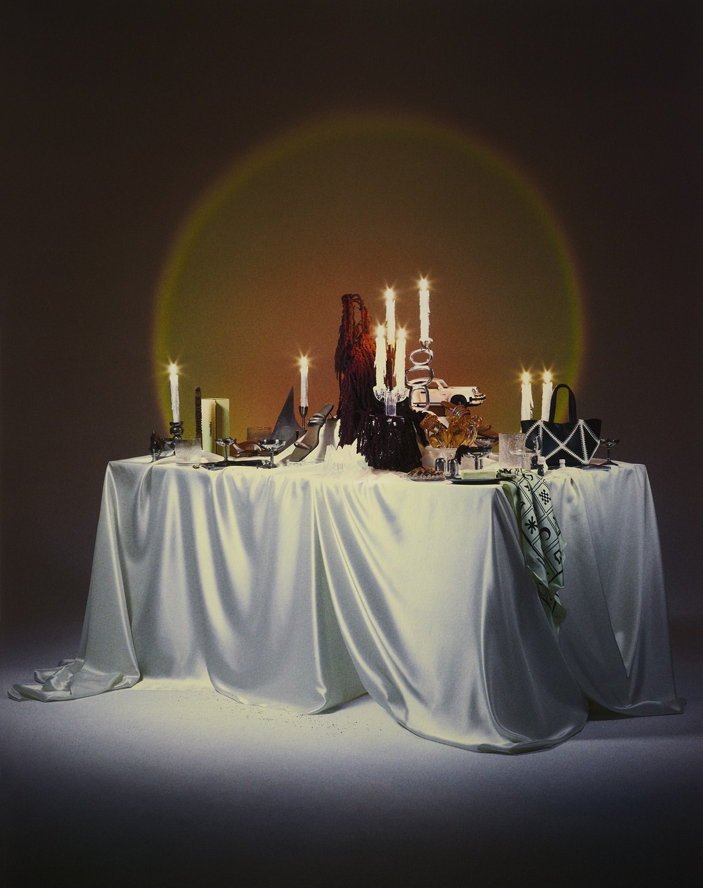 Indoor scene with a table draped in white fabric, adorned with candles, dishes, and decorative items, illuminated by a golden light source.