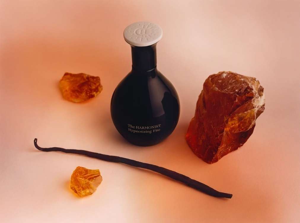 Still life shot of a black perfume bottle with a white cap, vanilla bean, and amber stones on a peach-colored backdrop, product photography.