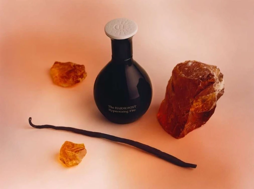 Still life shot of a black perfume bottle with a white cap, vanilla bean, and amber stones on a peach-colored backdrop, product photography.