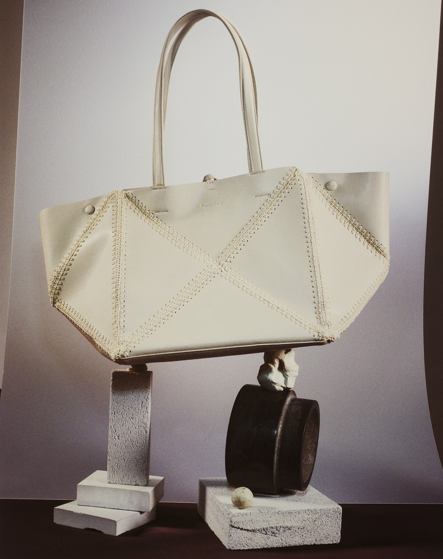 White leather tote bag with decorative stitching on a neutral background, styled on pedestals with geometric shapes and a dark container.