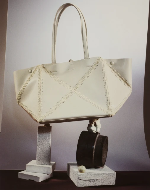 White leather tote bag with decorative stitching on a neutral background, styled on pedestals with geometric shapes and a dark container.