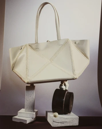 White leather tote bag with decorative stitching on a neutral background, styled on pedestals with geometric shapes and a dark container.