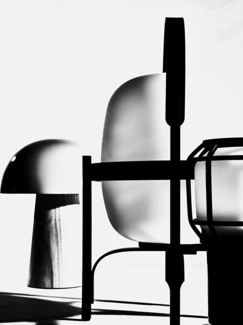 Monochrome photograph featuring a chair, a table lamp, and a cage-like object, rendered in high contrast with strong shadows on a light background.