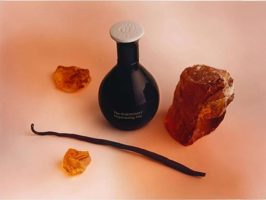 Still life shot of a perfume bottle, vanilla bean and amber stones on a peach background, showcasing beauty product and natural elements.